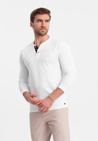 Weißes Langarm-Henley-Shirt mit schwarzen und goldenen Knöpfen am Kragen, kombiniert mit hellbeigen Hosen, die eine lässige Passform und eine glatte Textur aufweisen.