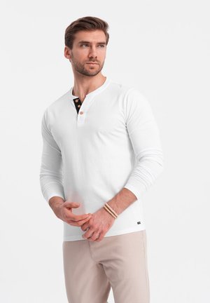 Camisa henley blanca de manga larga con botones negros y dorados en el cuello, combinada con pantalones beige claros, con un ajuste casual y una textura suave.