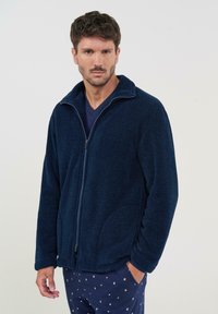 Chaqueta de forro polar azul marino con cremallera, cuello alto, tela texturizada y bolsillos laterales, llevada sobre una camisa oscura y pantalones de pijama con estampado.