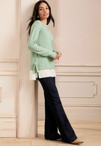 Hellgrüner Strickpullover mit Spitzenverzierungen an den Ärmeln, über einem weißen Hemd getragen. Kombiniert mit dunkelblauen Schlagjeans und hellbraunen High Heels.