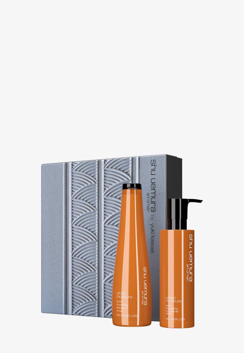 L'ensemble comprend deux bouteilles de shampooing et d'après-shampooing orange, accompagnées d'une boîte grise texturée présentant un motif de vague et des détails de logo.