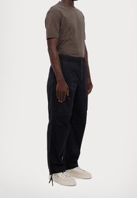 Brown cotton t-shirt with logo print, paired with black cargo pants featuring multiple pockets and elastic waistband, and light sneakers.
