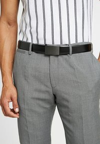 Pantalon gris ajusté avec une texture subtile, associé à une ceinture en cuir noir avec une boucle rectangulaire simple. Chemise à rayures verticales au-dessus.