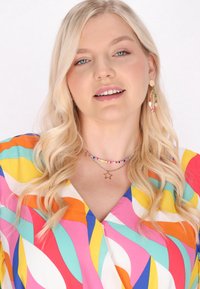 Rochie multicolor cu modele geometrice îndrăznețe, coliere suprapuse și cercei tip inel. Material moale cu o textură netedă și culori vibrante.