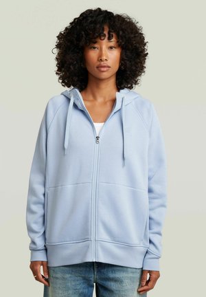 Woman with curly hair wearing a light blue zip-up hoodie and blue jeans, standing against a plain light background.