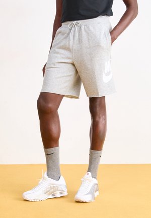 Persona che indossa pantaloncini grigi chiaro Nike, calzini grigi Nike, sneaker bianche Nike, in piedi su un pavimento giallo con le mani in tasca.