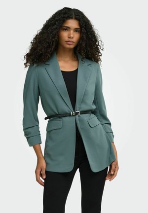 Kvinde med krøllet hår iført en teal blazer strammet ind med et tyndt sort bælte over en sort top og sorte bukser, stående foran en lys baggrund.