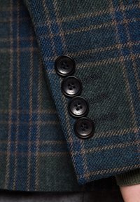 Rękaw marynarki w deseń w ciemnozieloną i niebieską kratę, z czterema czarnymi guzikami o teksturowanej powierzchni. Zbliżenie ukazuje detale tkaniny.