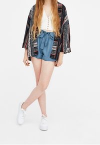 Pantaloni corti in tessuto blu con un dettagli di vita legata, abbinati a una maglietta corta bianca con bottoni e a un kimono stampato in tonalità scure.