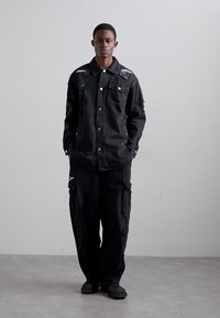 Jachetă din denim negru cu detalii uzate și nasturi tip snap, purtată împreună cu pantaloni cargo negri cu buzunare și o croială lejeră. Încălțăminte neagră.
