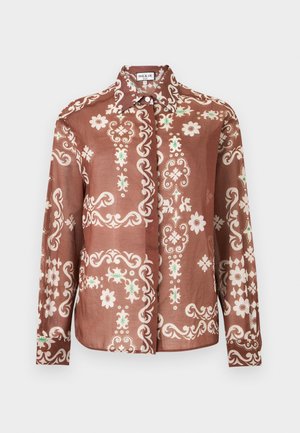 Femme portant un chemisier à motif floral couleur rouille rentré dans un pantalon large beige taille haute avec une ceinture, debout avec une main dans la poche.