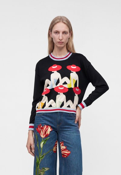 Femme portant un pull noir avec des figures au chapeau rouge et un jean bleu orné d'une grande broderie de fleur rouge sur une jambe.