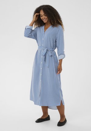 Kék-fehér csíkos ingruha, hosszú ujjakkal, gombos elejével, derékkötővel és oldalsó sliccekkel. Fekete lapos cipővel viselve.