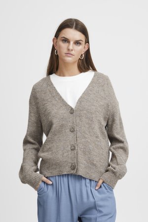 Lys brunt strikket cardigan med V-hals, med knappeåpning, lange ermer og ribbestrikkede mansjetter, brukt over en hvit T-skjorte.