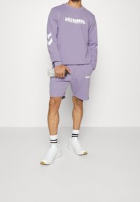 Fialová mikina s bílým logem, kombinovaná s odpovídajícími šortkami. Bílé sportovní boty a pruhované ponožky dokreslují celý outfit.