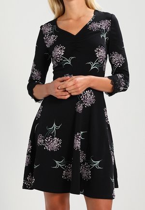 Vrouw draagt een zwarte V-hals jurk met paarse bloemmotieven en kanten afwerking op driekwart mouwen, handen in elkaar gevouwen ter hoogte van de taille.