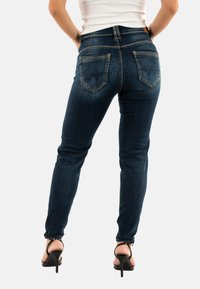 Jean skinny bleu foncé en denim, avec une taille mi-haute, des poches arrière et des coutures contrastées. Associé à des sandales à talons noirs.