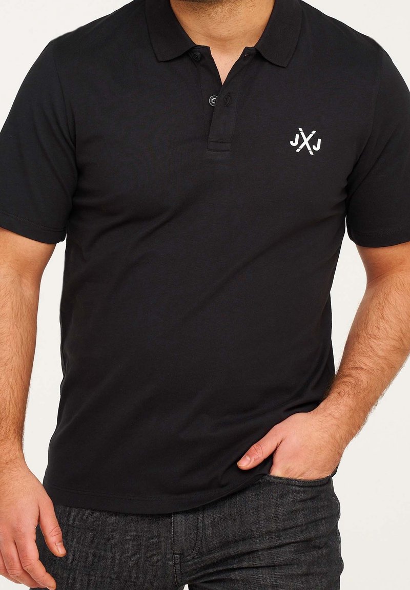 Homme portant un polo noir à manches courtes avec un petit logo blanc "JXJ", une main reposant dans la poche d'un jean foncé.