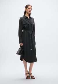 Μαύρο φόρεμα με μακριά μανίκια, κουμπιά μπροστά, δύο τσέπες στο στήθος και ζώνη στη μέση. Διαθέτει ίσια γραμμή με ασύμμετρο τελείωμα με ψηλότερο πίσω μέρος.