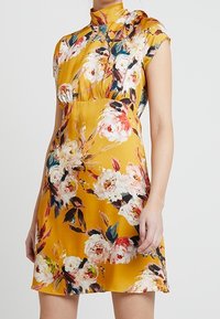 Robe florale en satin de couleur jaune moutarde avec des manches courtes et un col montant. Présente de grands motifs de roses en rose et des accents verts.