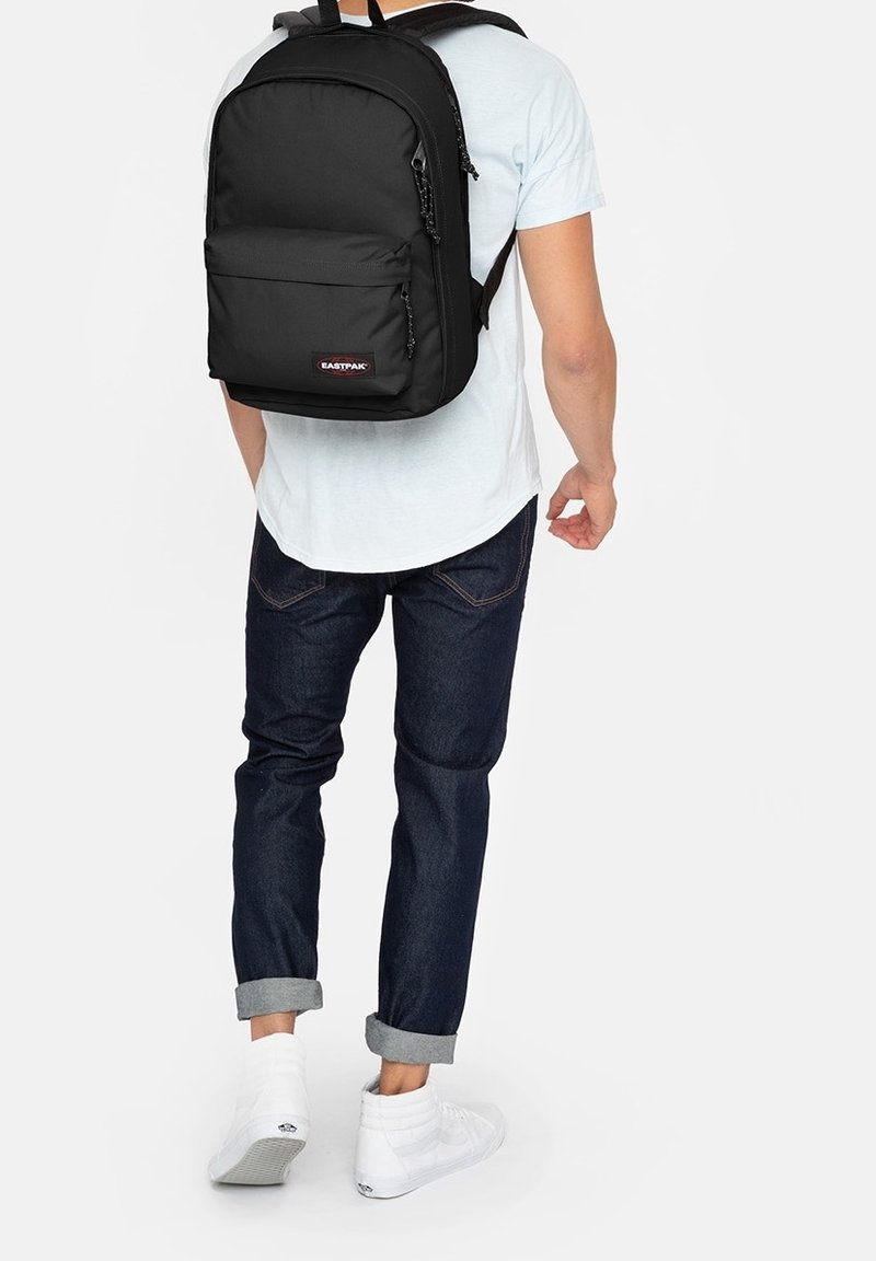 Černý batoh Eastpak se dvěma předními kapsami, polstrovanými popruhy a hladkým textilním povrchem, který nese osoba ve světle modrém tričku a džínech.