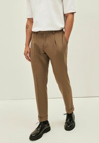 Pantaloni marroni su misura con pince frontali, realizzati in tessuto liscio. Abbinati a scarpe nere stringate. Camicia bianca semplice, in piedi su un pavimento strutturato.