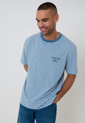 Jonge man glimlacht, draagt een blauw-wit gestreept T-shirt met blauwe spijkerbroek, staat met één hand in de zak tegen een effen achtergrond.