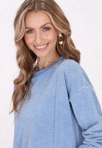 Felpa oversize azzurro chiaro con una consistenza morbida. Il modello ha lunghi capelli mossi e indossa collane a strati e orecchini colorati.