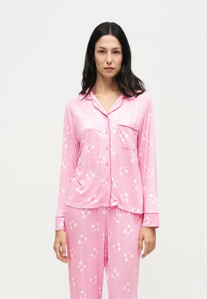 Femme aux longs cheveux foncés portant un pyjama en satin rose avec des motifs de nœuds blancs, debout devant un fond clair uni.