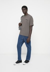 T-shirt gris à manches courtes avec détail de logo, associé à un jean en denim bleu et des sneakers noires. Le modèle porte un sac à bandoulière gris.
