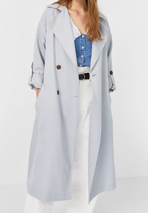 Femme portant un long trench-coat gris clair par-dessus un haut bleu à boutons et un pantalon taille haute blanc avec ceinture noire, mains dans les poches.