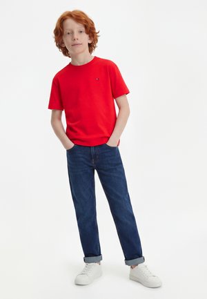 Ragazzo dai capelli rossi che indossa una maglietta rossa, jeans blu con risvolti, scarpe da ginnastica bianche, in piedi con le mani nelle tasche su sfondo bianco.