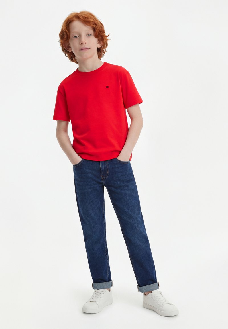 Αγόρι με κόκκινα μαλλιά που φοράει κόκκινη μπλούζα, μπλε τζιν με γυρισμένο τελείωμα, λευκά αθλητικά παπούτσια, στέκεται με τα χέρια στις τσέπες σε λευκό φόντο.