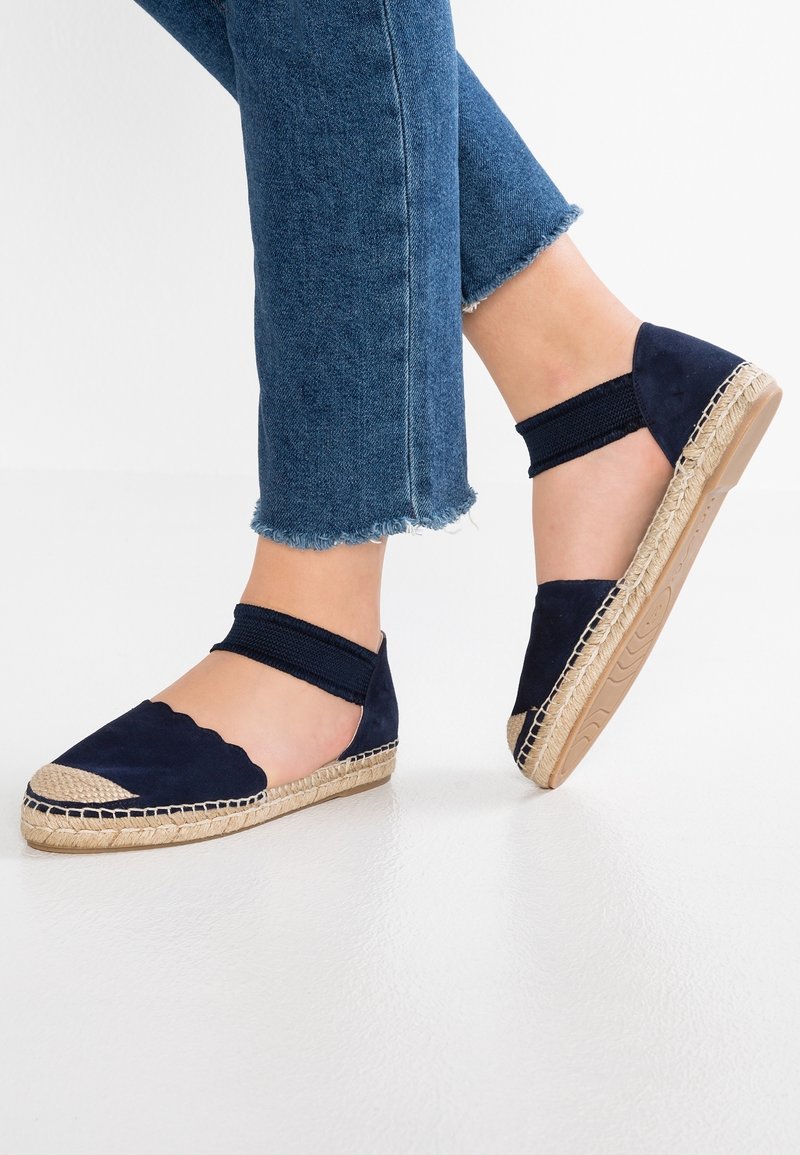 Pieds portant des espadrilles plates bleu marine avec bouts tissés et brides de cheville, associées à un jean bleu raccourci sur un sol blanc.