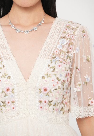 Φόρεμα σε εκρού απόχρωση με κεντήματα λουλουδιών, λεπτομέρειες από δαντέλα και διαφανείς κοντές μανσέτες. Συνοδεύεται από ασημένιο λουλουδένιο κολιέ.