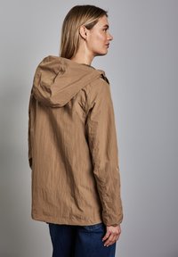 Světle hnědá, lehká bunda s kapucí, texturovaná tkanina, volný střih, viditelné švy a jednoduchý design. Nošena přes tmavě modré džíny.