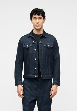 Νεαρός άνδρας στέκεται πρόσωπο προς τα εμπρός, φοράει σκούρο τζιν μπουφάν με κουμπωτές τσέπες στο στήθος και σκουρόχρωμο ριγέ παντελόνι.