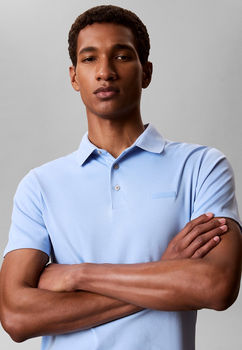 Jonge man met kort krullend haar, draagt een lichtblauw poloshirt, staat met gekruiste armen tegen een effen grijze achtergrond.