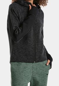 Sudadera con cremallera en negro y gris, con un acabado texturizado y mangas raglán. Combinada con pantalones jogger verdes, que cuentan con una cinturilla ajustable y un patrón sutil.