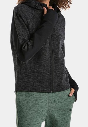 Sudadera con cremallera en negro y gris, con un acabado texturizado y mangas raglán. Combinada con pantalones jogger verdes, que cuentan con una cinturilla ajustable y un patrón sutil.