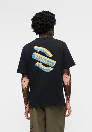 Person mit Vitiligo, trägt ein schwarzes T-Shirt mit einem blauen und gelben 3D-"Dickies"-Logo und Pfeilen auf der Rückseite, kombiniert mit olivfarbenen Hosen.