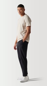 Camiseta beige con detalle de logo combinada con pantalones deportivos negros con bolsillos con cremallera, acompañada de zapatillas blancas. Silueta casual y ajustada.