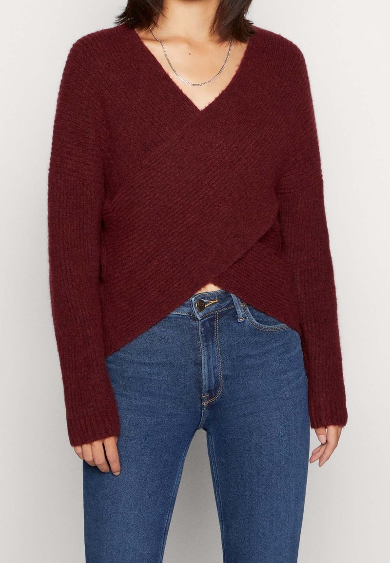 Femme portant un pull bordeaux texturé à col en V avec un devant croisé, associé à un jean bleu taille haute sur un fond uni.