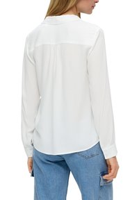 Camicia bianca a maniche lunghe con colletto, caratterizzata da una piega sul retro, texture liscia e polsini con bottoni, abbinata a jeans blu chiaro.
