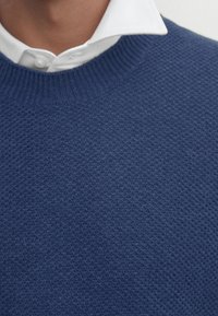 Pull en maille bleu marine avec une surface texturée et un col côtelé, porté sur une chemise blanche à col, présentant un design classique.