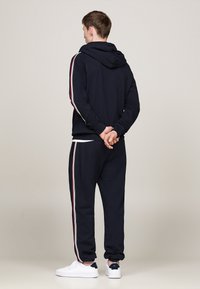 Chándal navy con capucha, con franjas laterales en blanco y rojo. Textura suave, puños elásticos, combinado con zapatillas blancas.