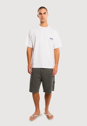 Mężczyzna w białej, luźnej koszulce, ciemnoszarych szortach za kolano oraz białych klapkach, stojący na tle jasnego, jednolitego tła.