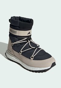 Beige a černá zateplená bota s elastickými tkaničkami a nastavitelným páskem kolem kotníku. Texturovaná podešev s výrazným vzorem. Značka "MOON BOOT" je umístěna na botě.