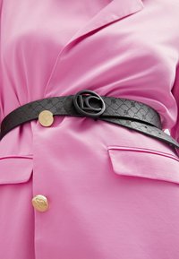 Svart mönstrad bälte med en rund logo-spänne, bärs över en rosa blazer med slät textur och guldknapplister.