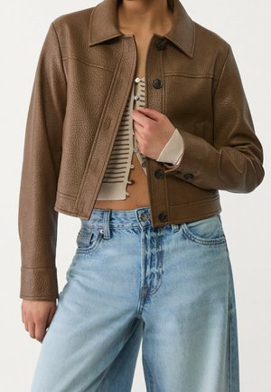 Person iført en cropped brun tekstureret læderjakke over en stribet, knappet crop top og lyseblå højtaljede jeans.